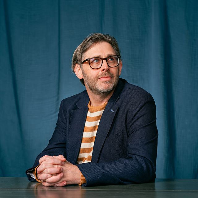 Man in a navy sport coat sits with hands folded in front of a blue curtain background